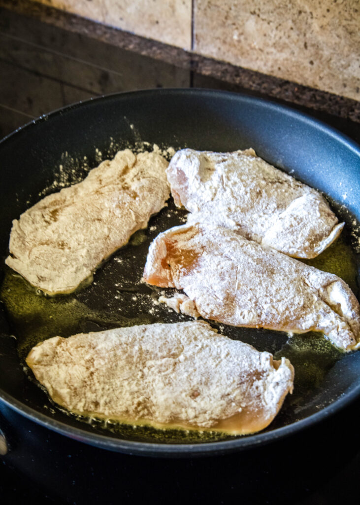 Four chicken breasts coated in seasonings and flour cooking in a skillet
