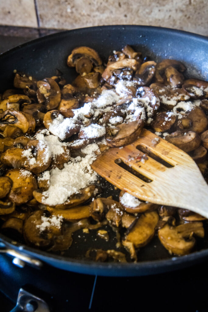 A wooden spatula stirring flour and mushrooms in a skillet