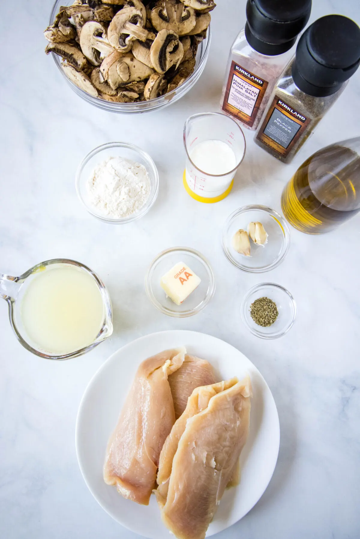 Overhead view of the ingredients needed for mushroom chicken: a plate of chicken breasts, a pyrex of chicken broth, a bowl of mushrooms, a bottle of olive oil, a glass of cream, a knob of butter, a bowl of flour, a bowl of garlic, a bowl of Italian seasoning, and salt and pepper grinders