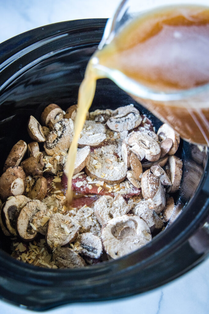 pouring broth into crockpot with mushrooms and cube steak