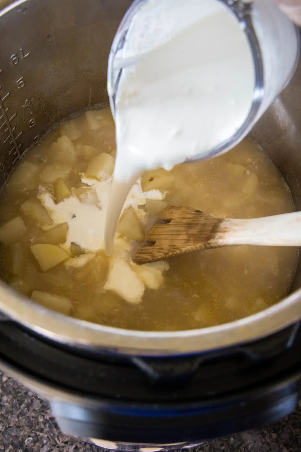 A pyrex pouring cream into a pot of potato soup with a wooden spoon.