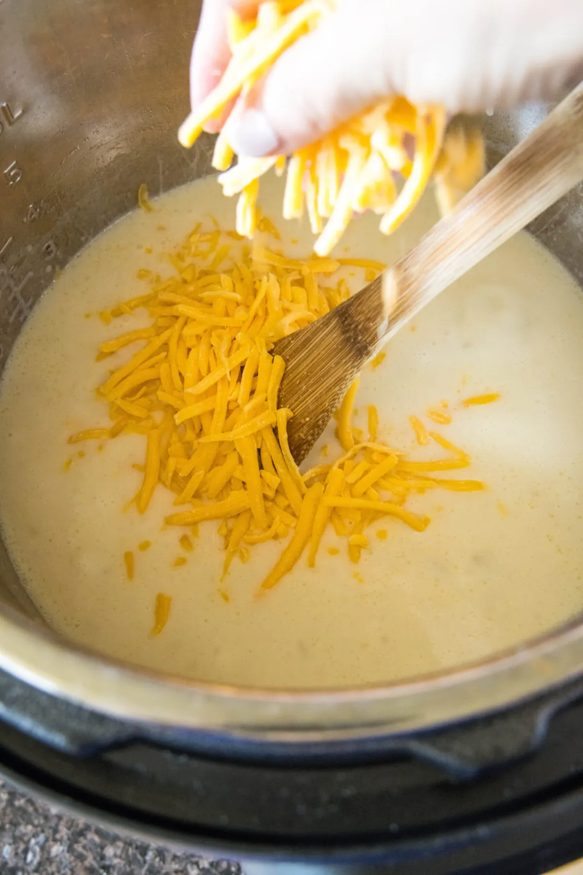 A hand dropping shredded cheese into a pot of potato soup with a wooden spoon.
