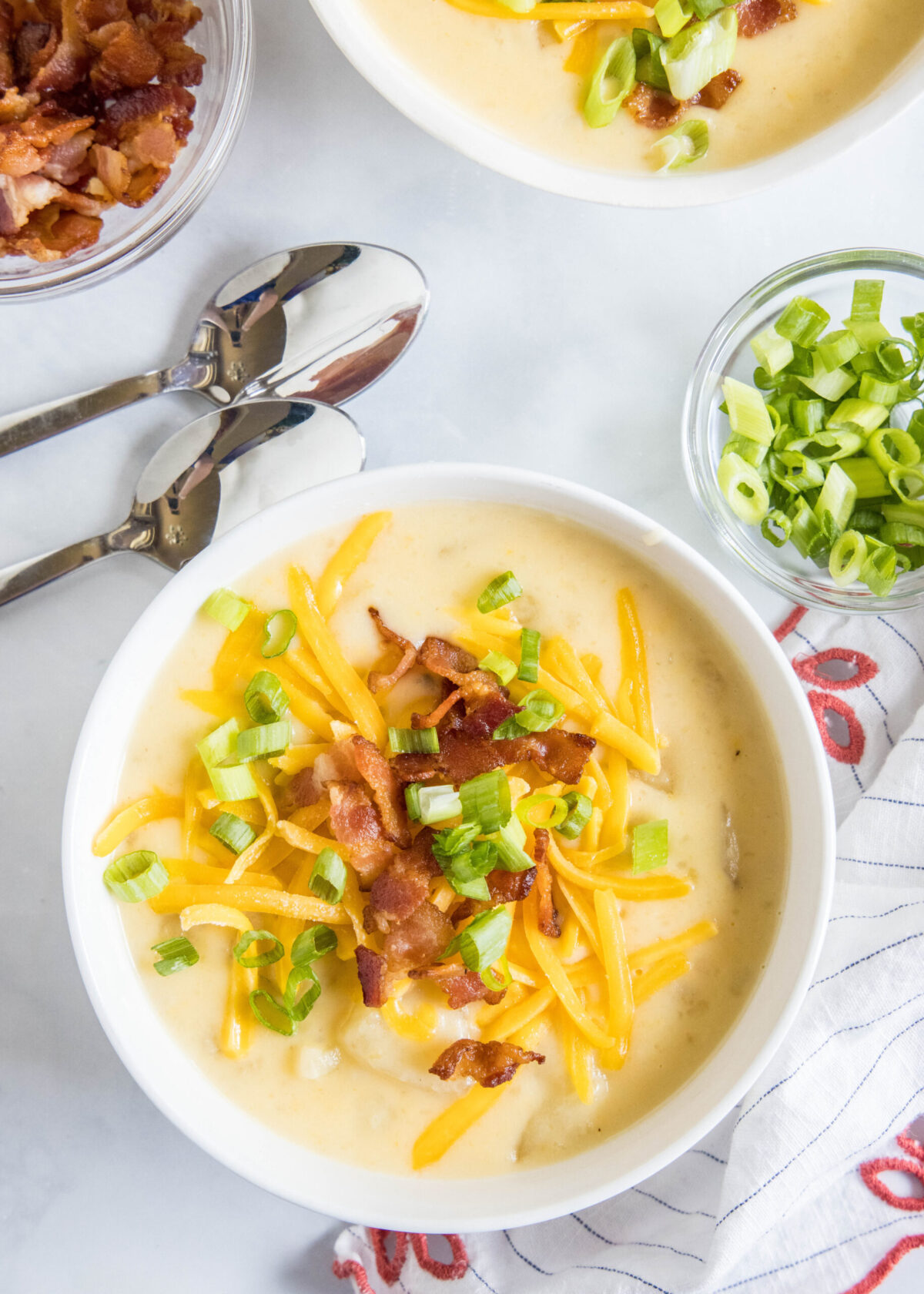 Overhead view of a bowl of potato soup topped with cheese, bacon, and green onions, next to two spoons, a bowl of green onions, a bowl of bacon, and another bowl of soup.