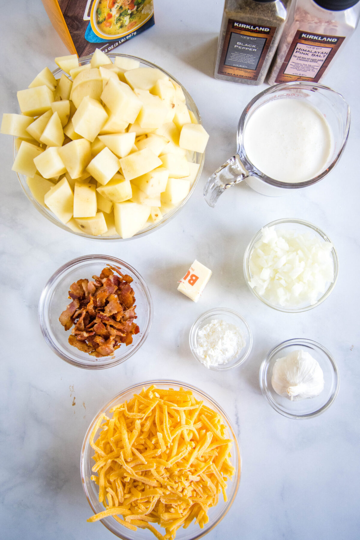 Overhead view of the ingredients needed for Instant Pot potato soup: a bowl of chopped potatoes, a bowl of shredded cheese, a bowl of crumbled bacon, a pyrex of cream, a bowl of onions, a bowl of garlic, a bowl of cornstarch, som butter, a carton of chicken stock, and salt and pepper grinders.