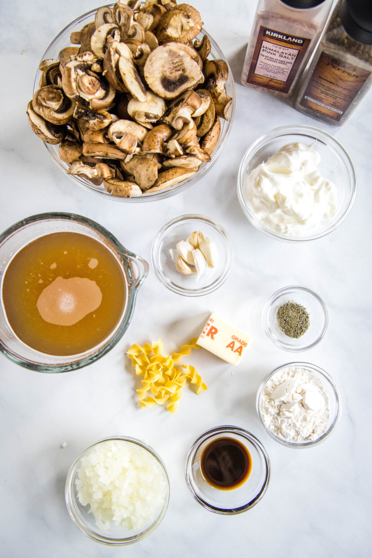 Overhead view of the ingredients needed for mushroom stroganoff: a bowl of sliced mushrooms, a pyrex of broth, a bowl of sour cream, a bowl of garlic, a bowl of flour, a bowl of onions, a bowl of Worcestershire sauce, half a stick of butter, a bowl of thyme, some dried egg noodles, and a salt and pepper grinder