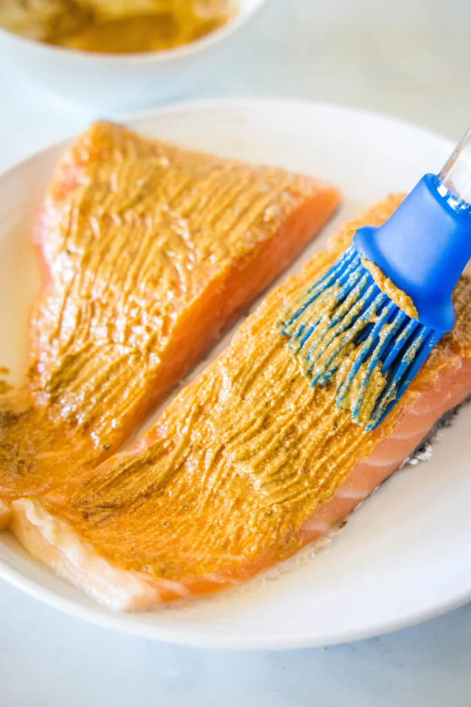 A brush rubbing a mustard spread on two raw salmon fillets.