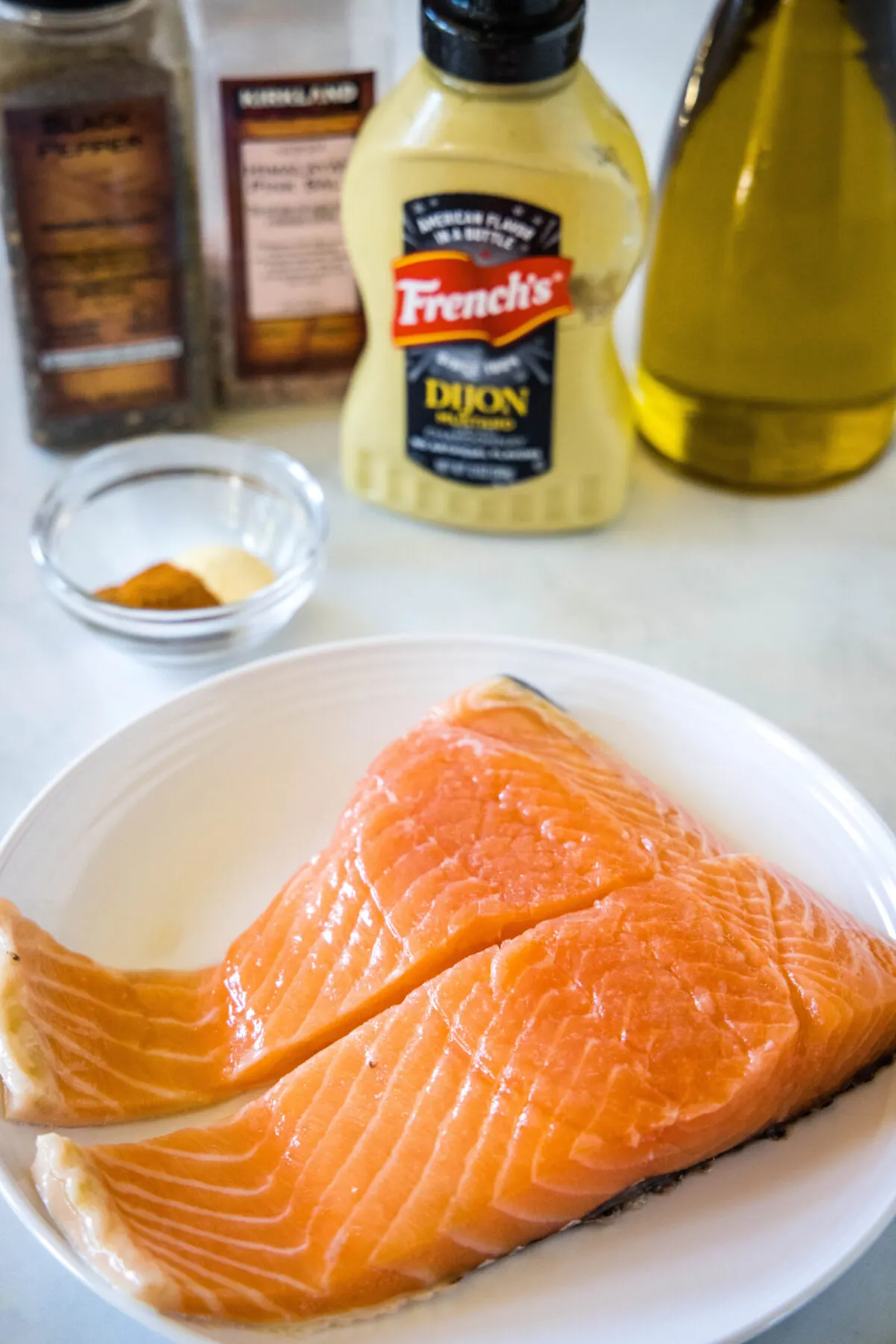 All the ingredients needed to make air fryer salmon: a plate with two salmon fillets, a bowl with garlic powder and paprika, a bottle of Dijon mustard, a bottle of olive oil, and salt and pepper grinders.