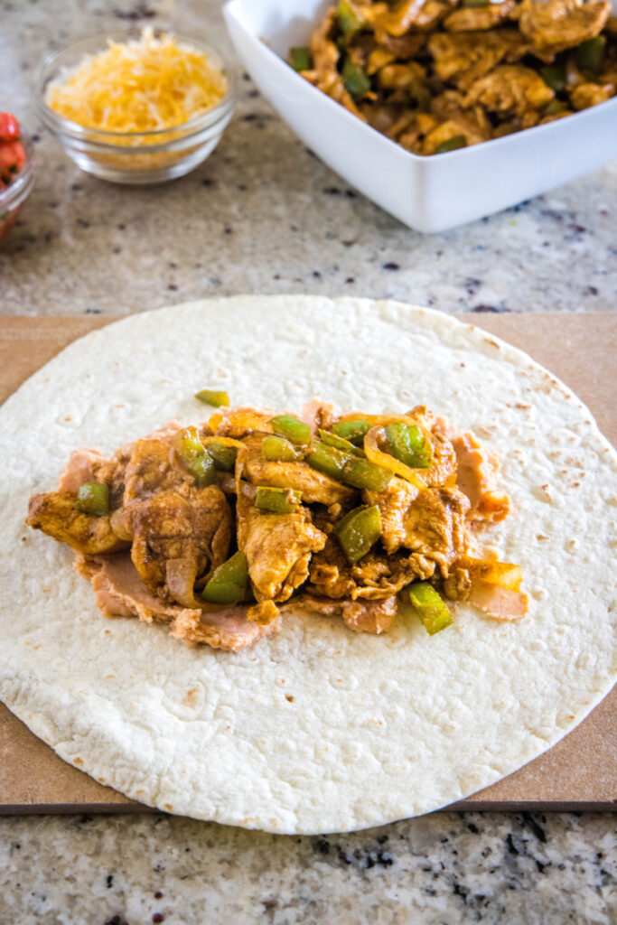A tortilla topped with beans and chicken fajita mix, next to a bowl of chicken fajitas and a bowl of cheese.