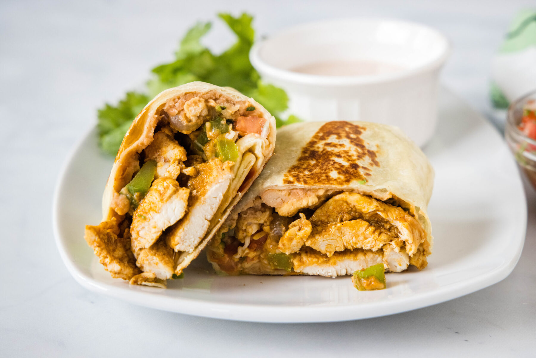 A plate with two halves of a chicken fajita wrap, with a bowl of sauce and lettuce in the background.
