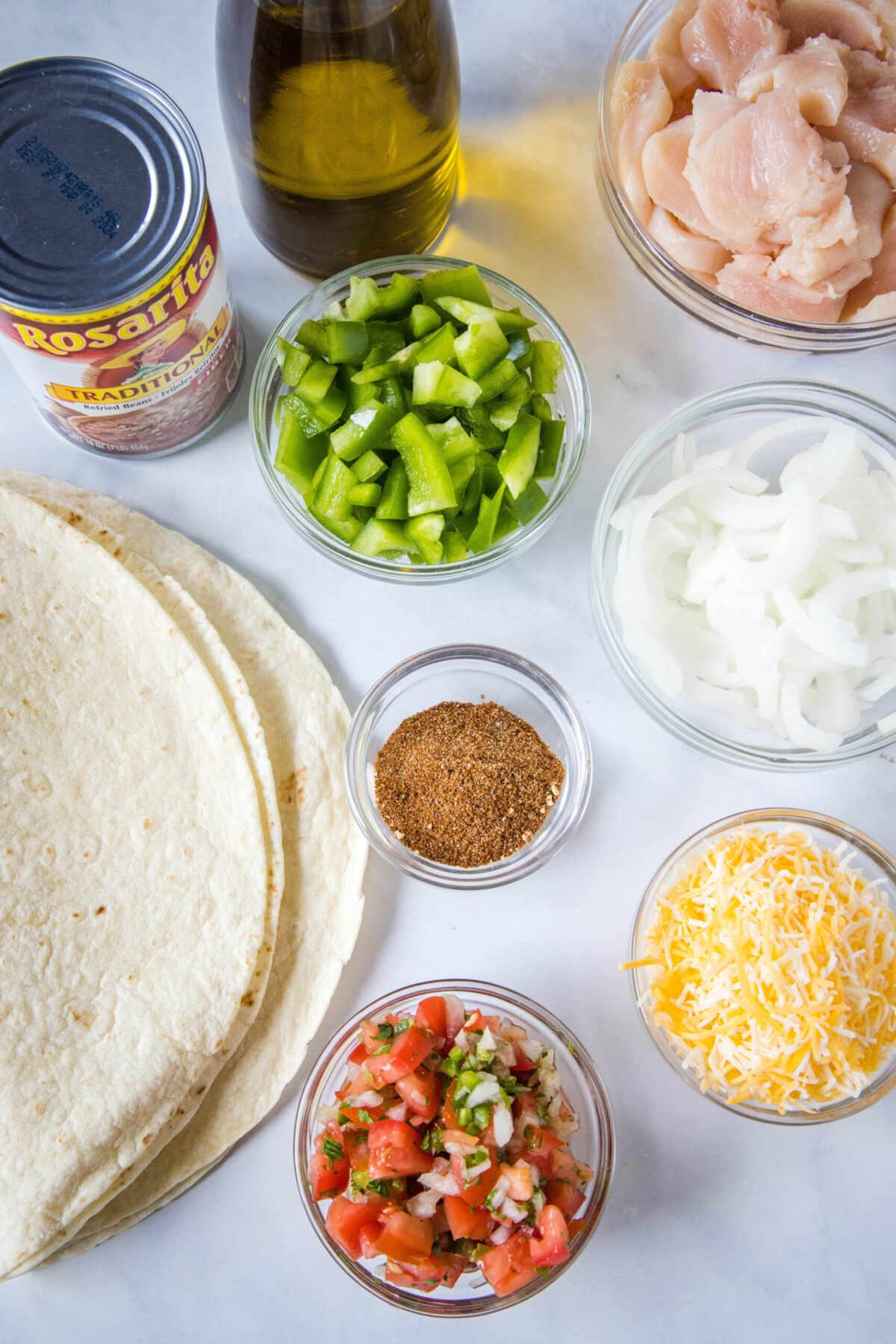 Overhead view of the ingredients needed for chicken fajita wraps: a stack of tortillas, a can of refried beans, a bottle of olive oil, a bowl of chicken, a bowl of onions, a bowl of green peppers, a bowl of shredded cheese, a bowl of pico de gallo, and a bowl of spices.