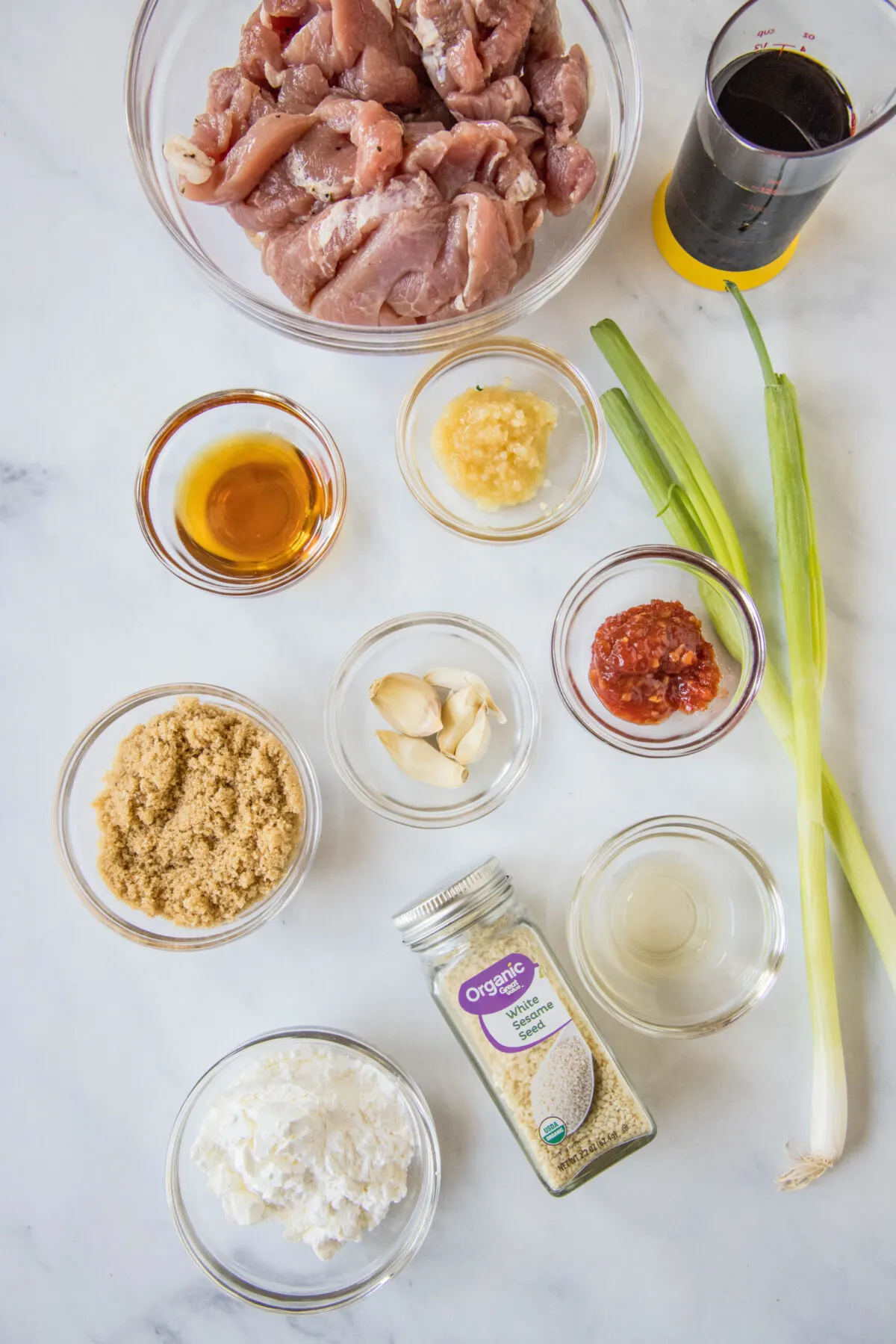 Overhead view of the ingredients needed for Mongolian pork: a bowl of pork strips, a glass of soy sauce, a glass of vinegar, a bowl of ginger, a bowl of garlic, a bowl of cornstarch, a bowl of brown sugar, a bowl of sesame oil, a jar of sesame seeds, a bowl of chili paste, and two green onions.