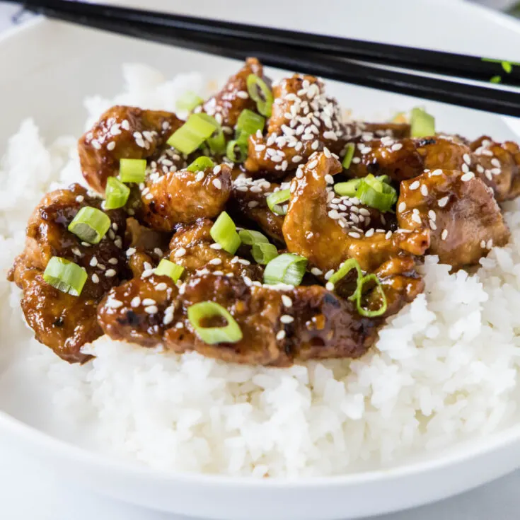 A plate with Mongolian pork on top of rice, topped with sesame seeds and green onions, with a pair of chopsticks.