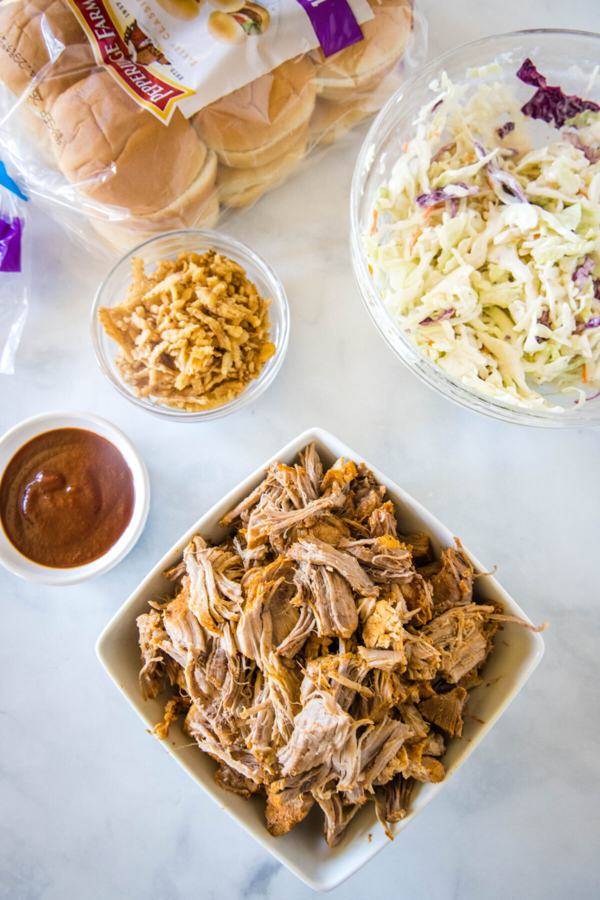 Overhead view of the ingredients needed for pulled pork sliders: a bowl of pulled pork, a bowl of coleslaw, a bowl of barbecue sauce, a bowl of fried onions, and a bag of slider buns.