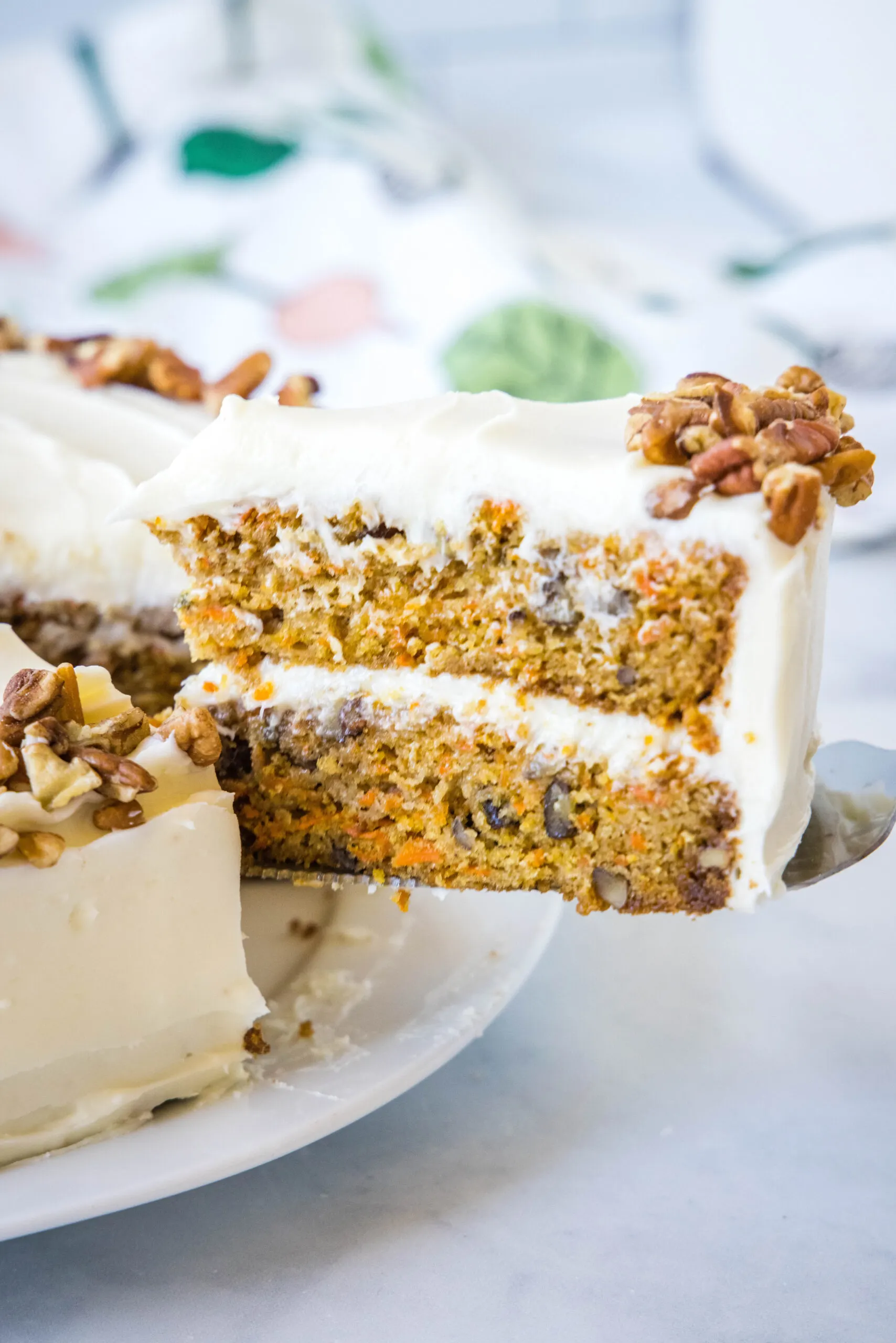 A slice of frosted carrot cake is lifted from the rest of the cake with a cake server.