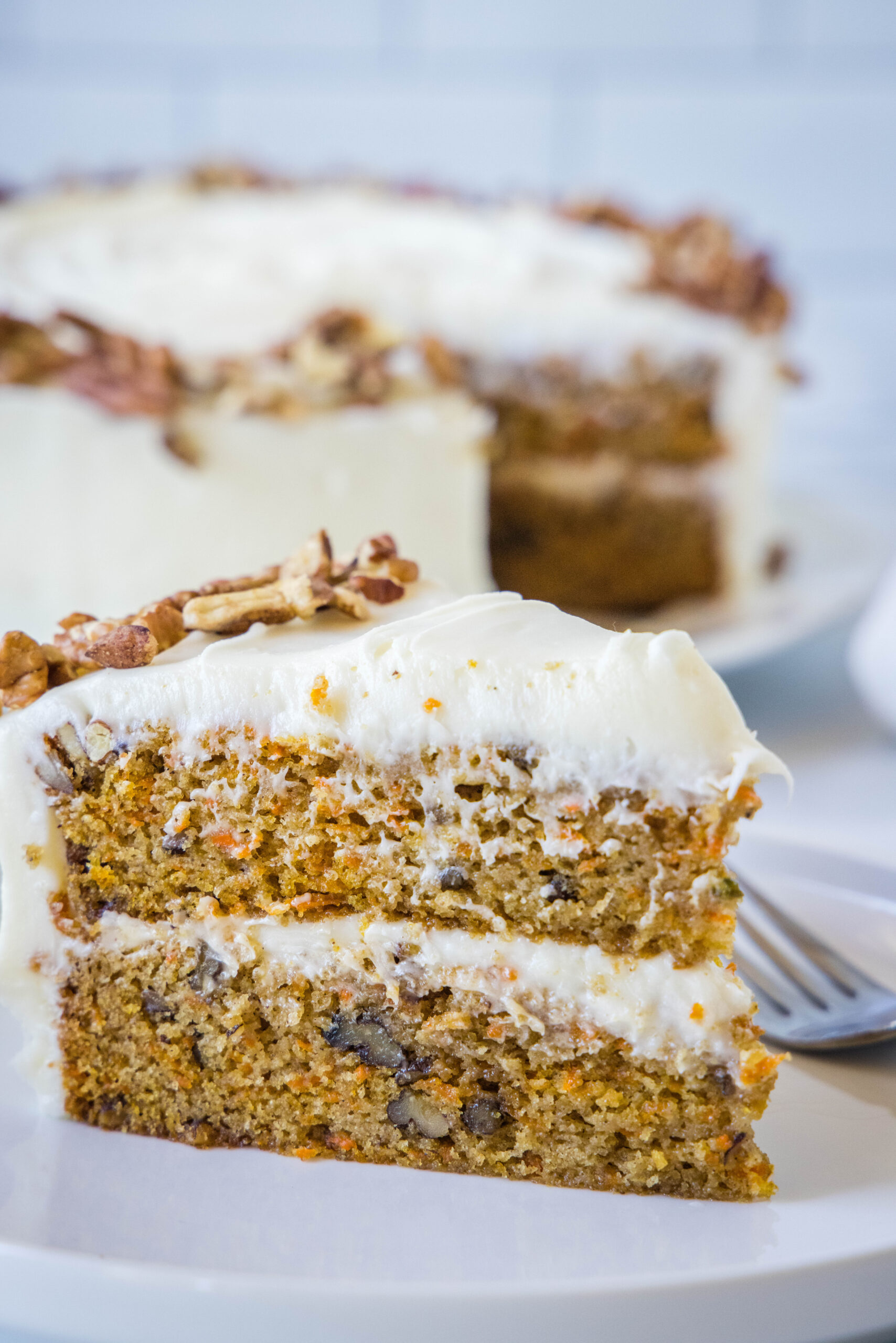 A slice of frosted carrot cake on a white plate next to a fork, with the rest of the cake in the background.