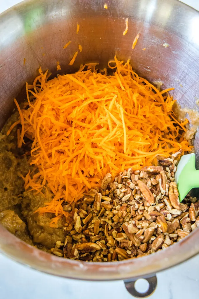 Shredded carrots and chopped pecans added to carrot cake batter in a metal mixing bowl.