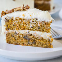 A slice of frosted carrot cake on a white plate next to a fork.