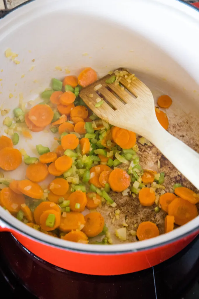 Sliced carrots, onion, and celery sauteing in a large pot with a wooden spatula.