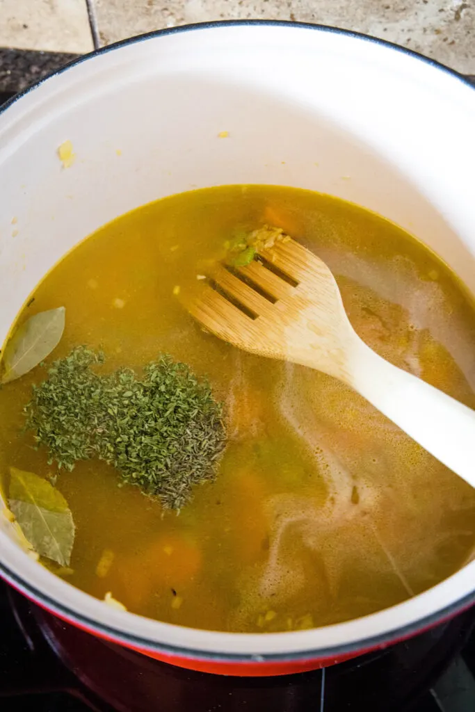 Chicken broth and dried herbs added to a pot with sauteed vegetables. with a wooden spatula.