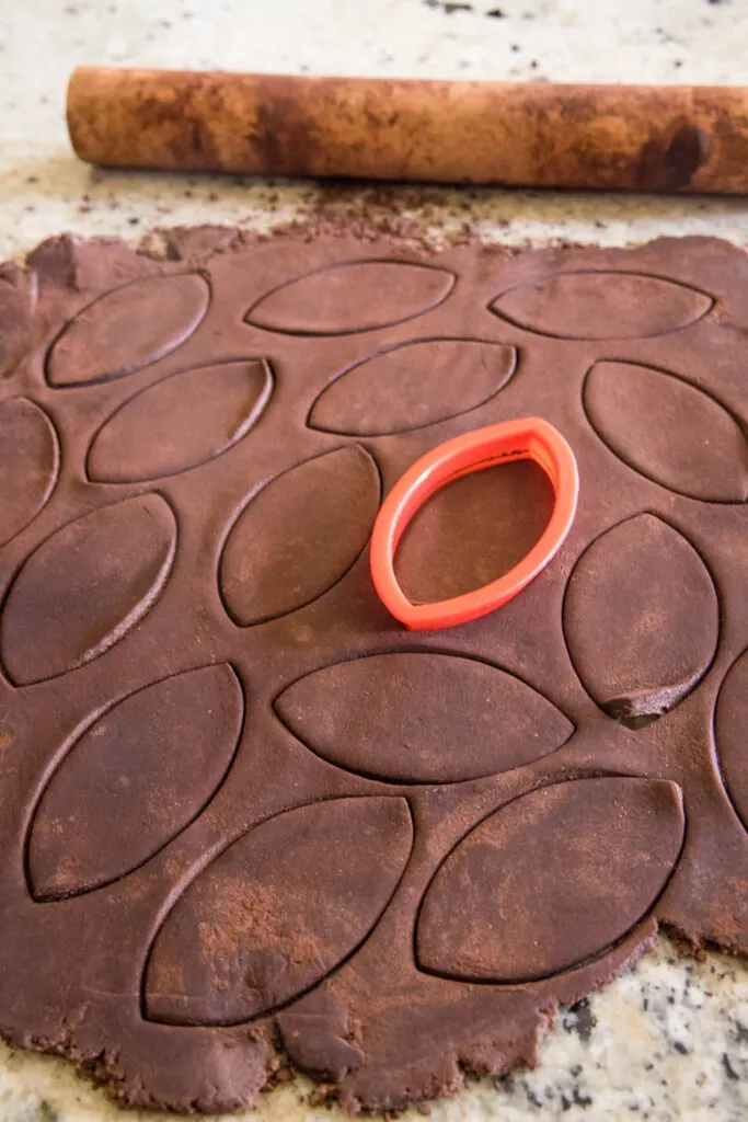 A red cookie cutter is used to cut chocolate sugar cookie dough into football shapes.