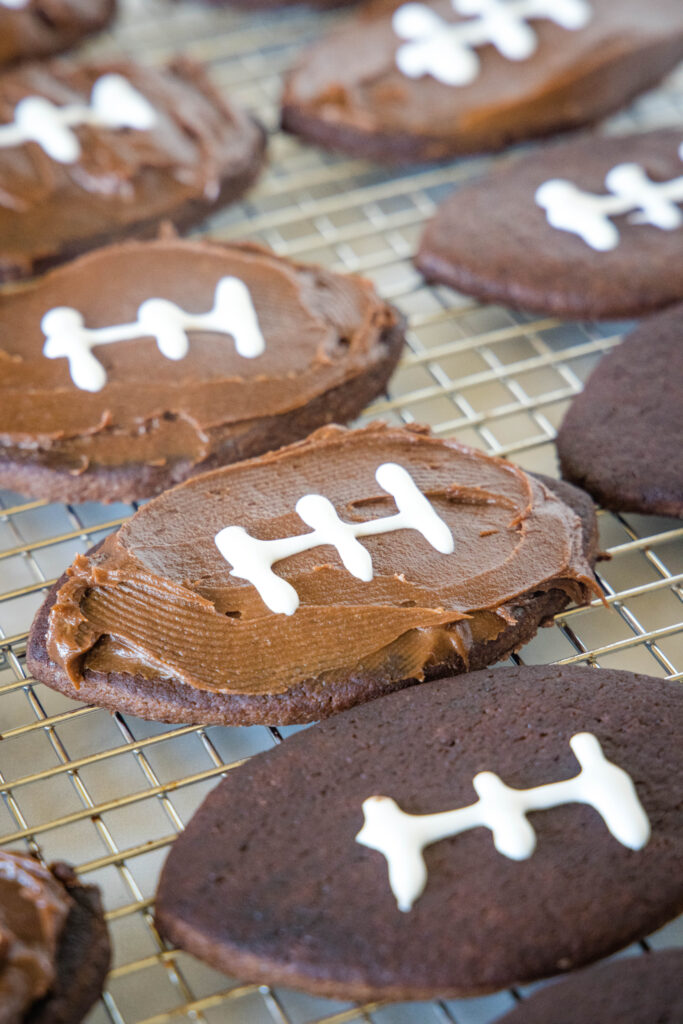 Chocolate sugar cookies partially frosted and decorated to look like footballs on a wire rack.