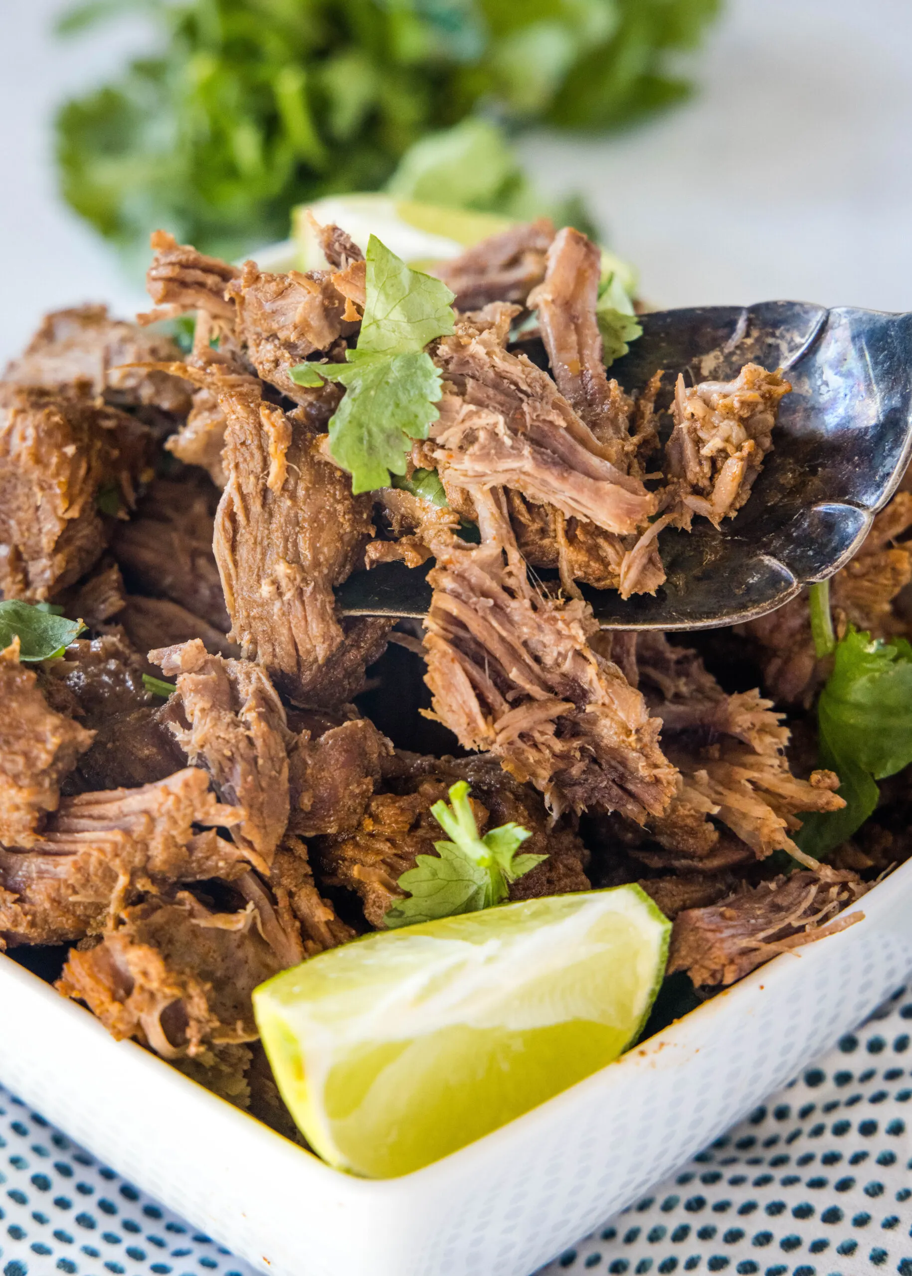 A serving spoon lifting Mexican shredded beef from a square bowl of beef garnished with cilantro and a lime wedge.