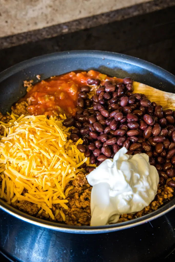 Salsa, black beans, sour cream, and shredded cheese added to a skillet with browned taco meat.