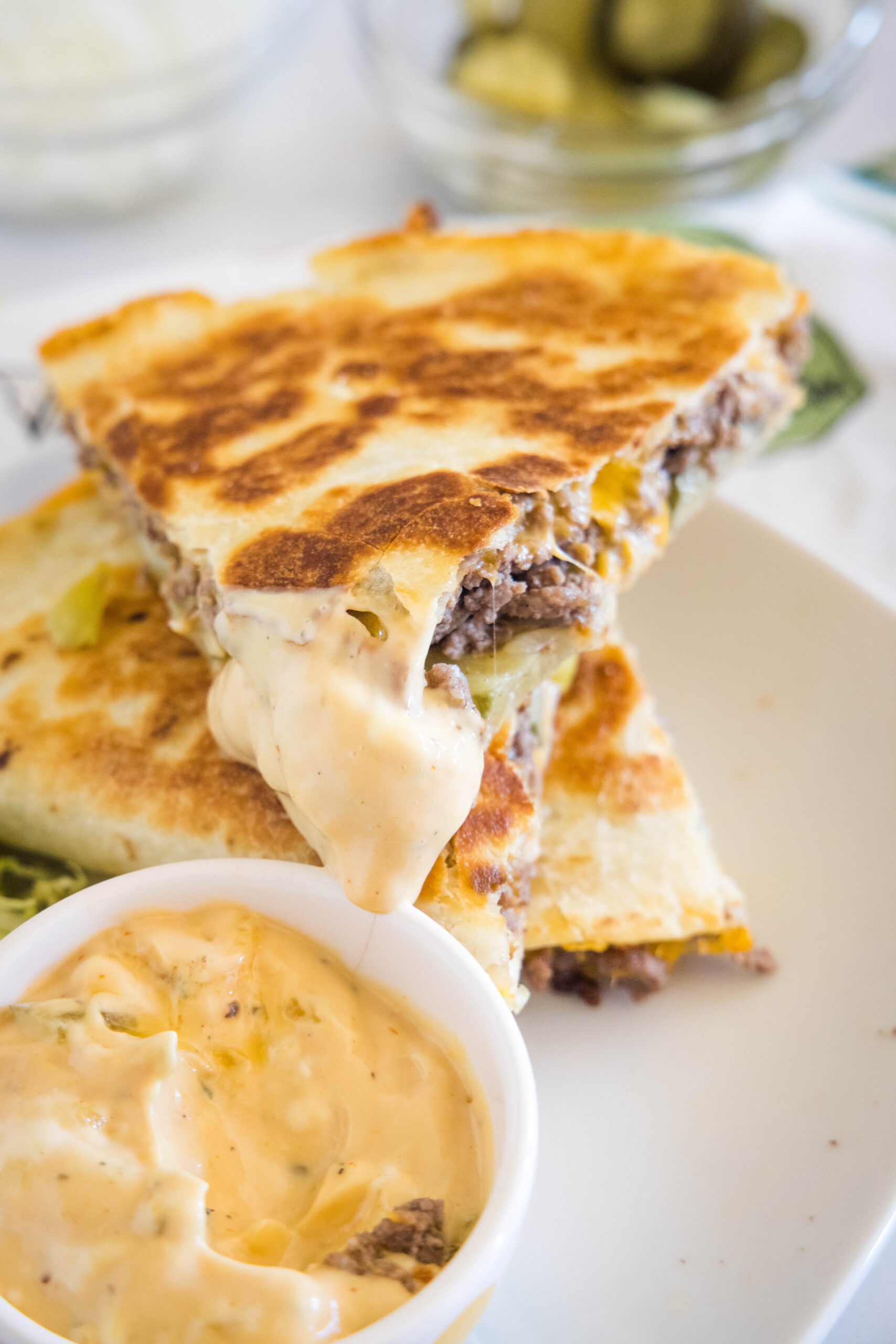 A slice of cheeseburger quesadilla dipped into a bowl of burger sauce, stacked on top of another slice on a white plate.