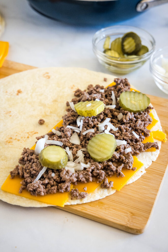 A flour tortilla laying open on a wooden board, one half topped with cheese, burger meat, onions, and pickles.