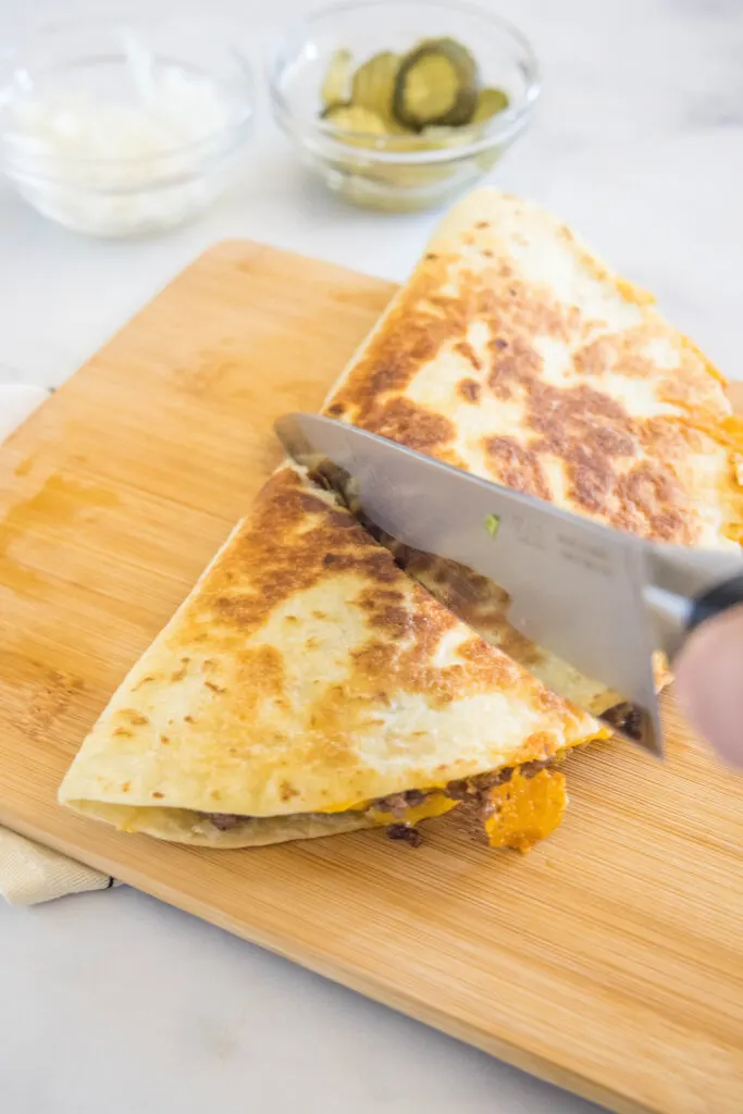 A knife cutting a cheeseburger quesadilla into slices on a wooden cutting board.