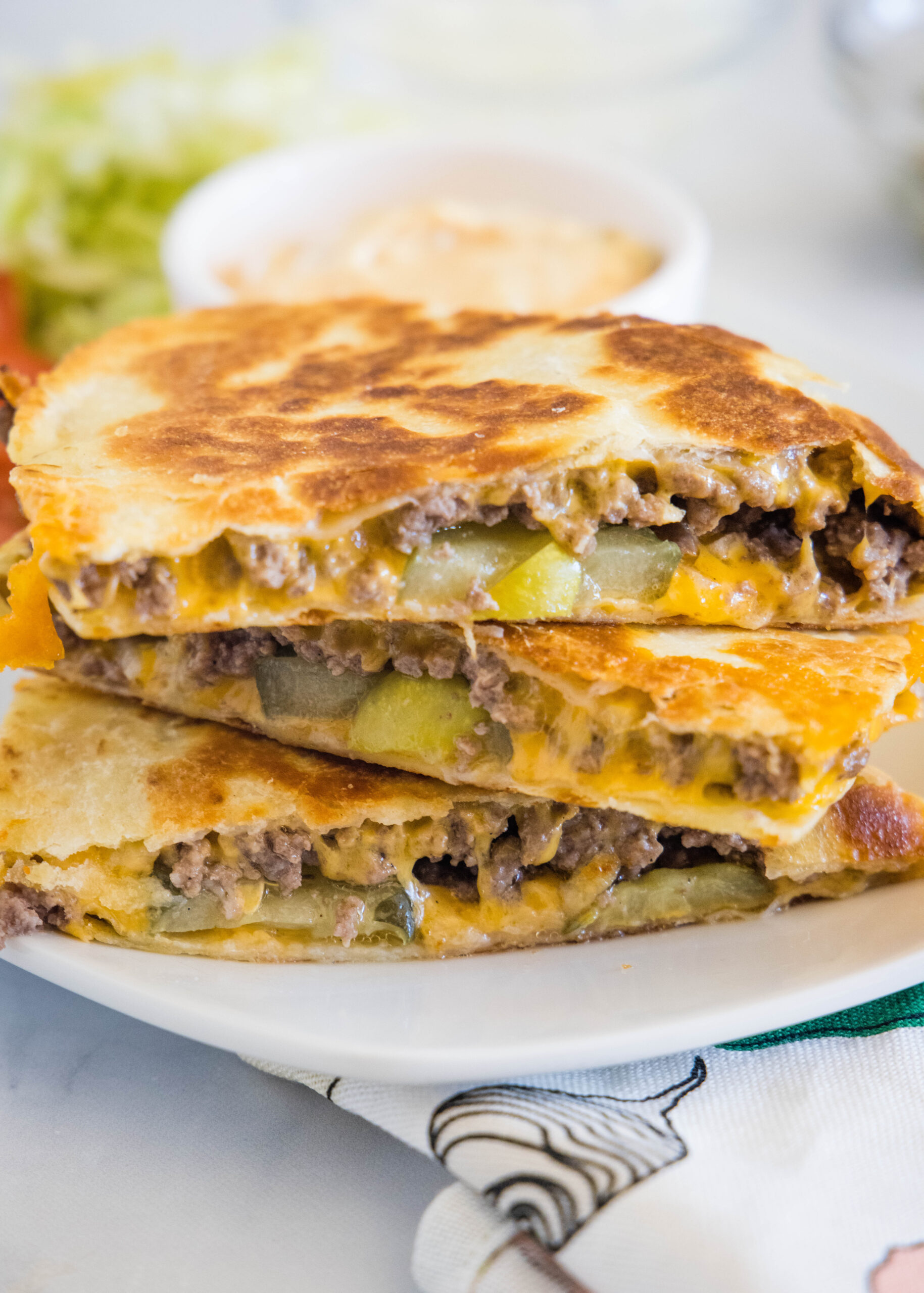 Three slices of cheeseburger quesadilla stacked on a plate, with a bowl of burger sauce in the background.