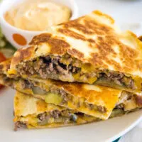 Three slices of cheeseburger quesadilla stacked on a plate, next to a bowl of burger sauce.