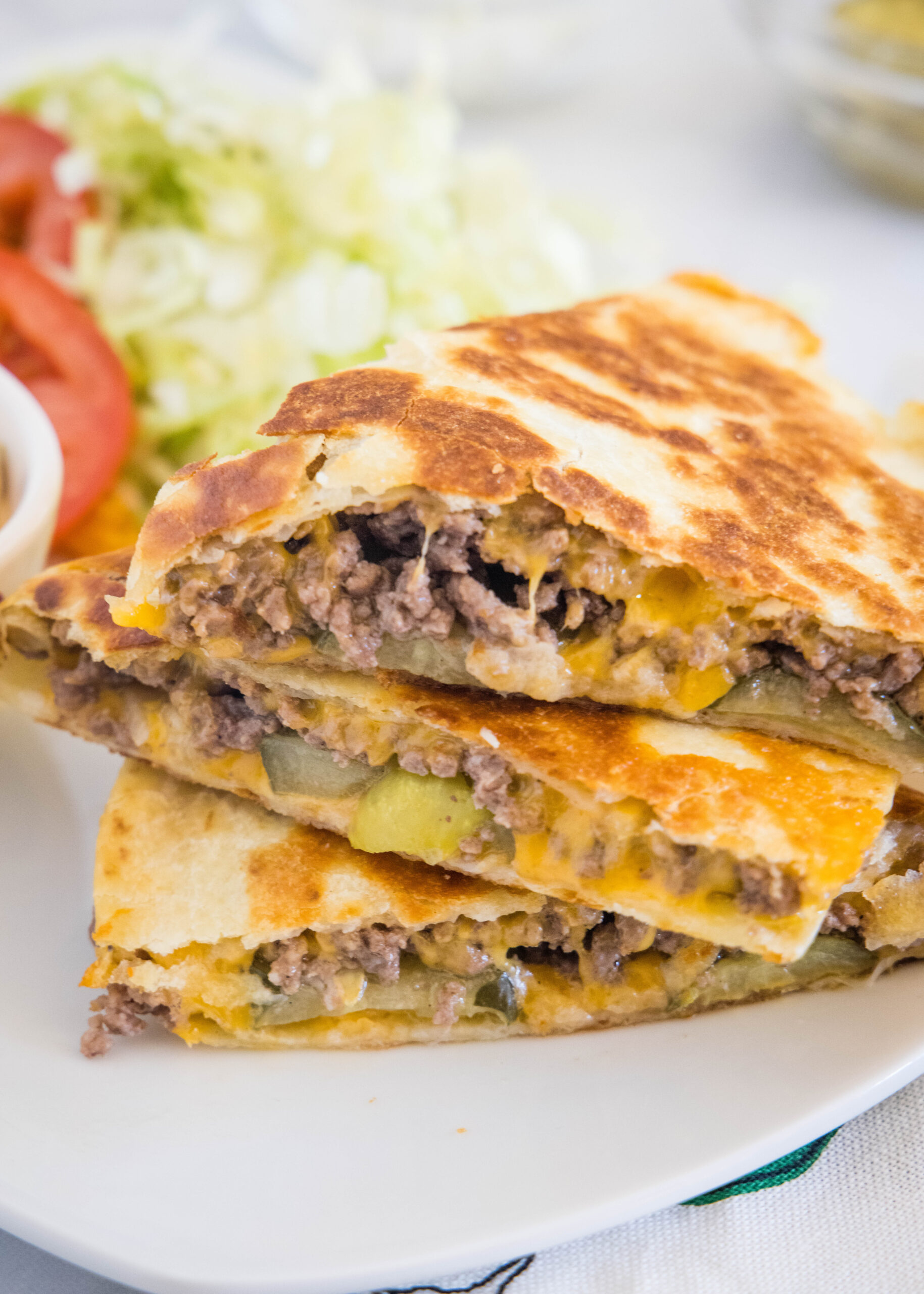 Three slices of cheeseburger quesadilla stacked on a plate, with lettuce and sliced tomatoes in the background.