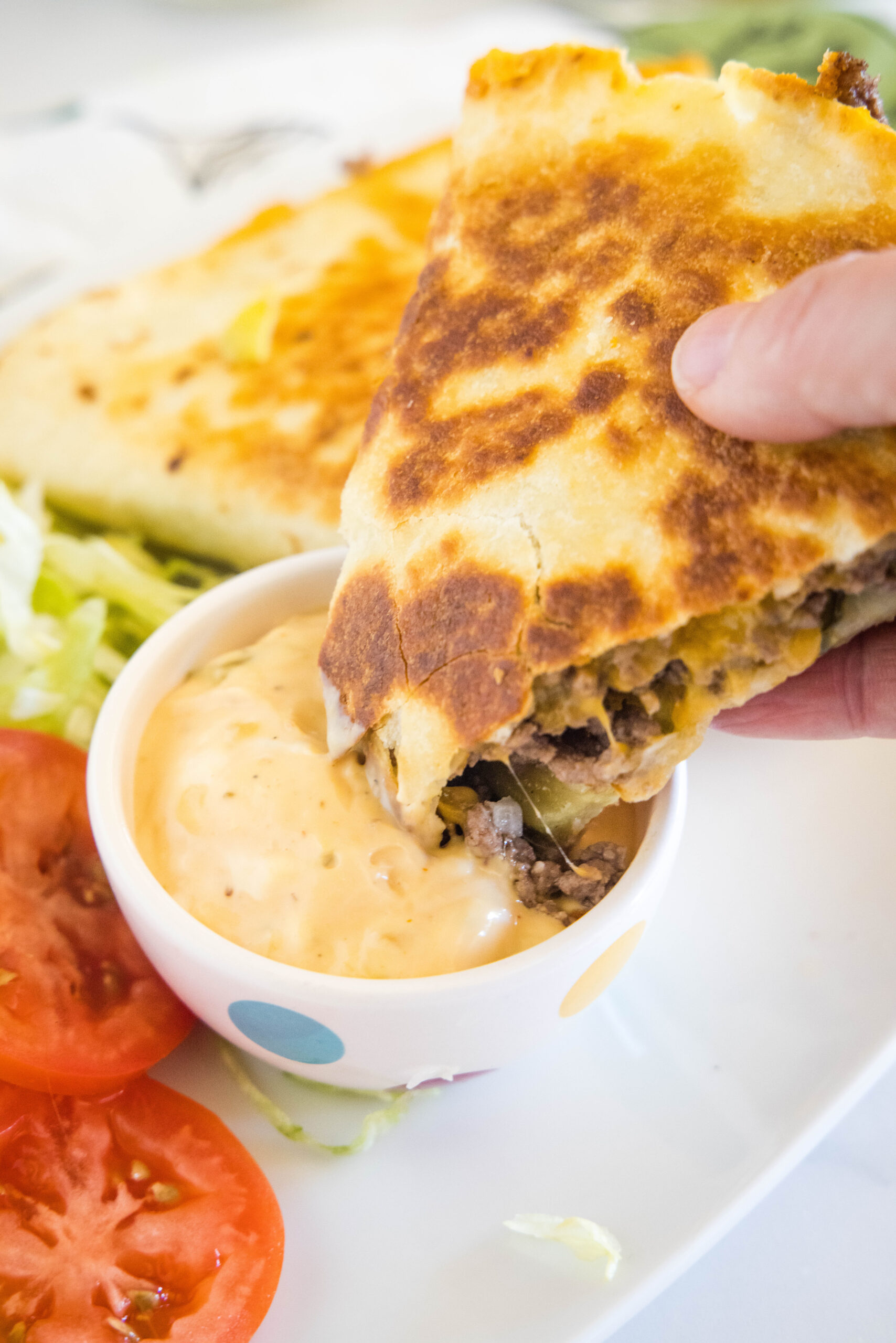 A hand dipping a cheeseburger quesadilla into a small bowl of burger sauce, with more quesadillas in the background.