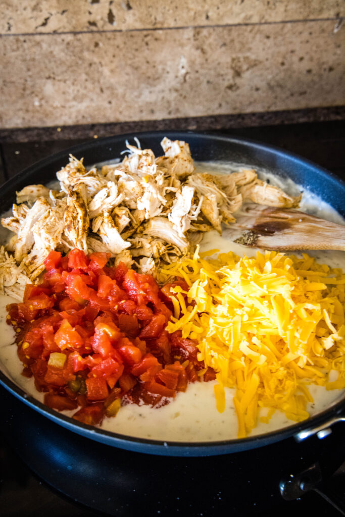 Rotel tomatoes, shredded chicken, and shredded cheese added to cream sauce in a skillet, next to a wooden spoon.