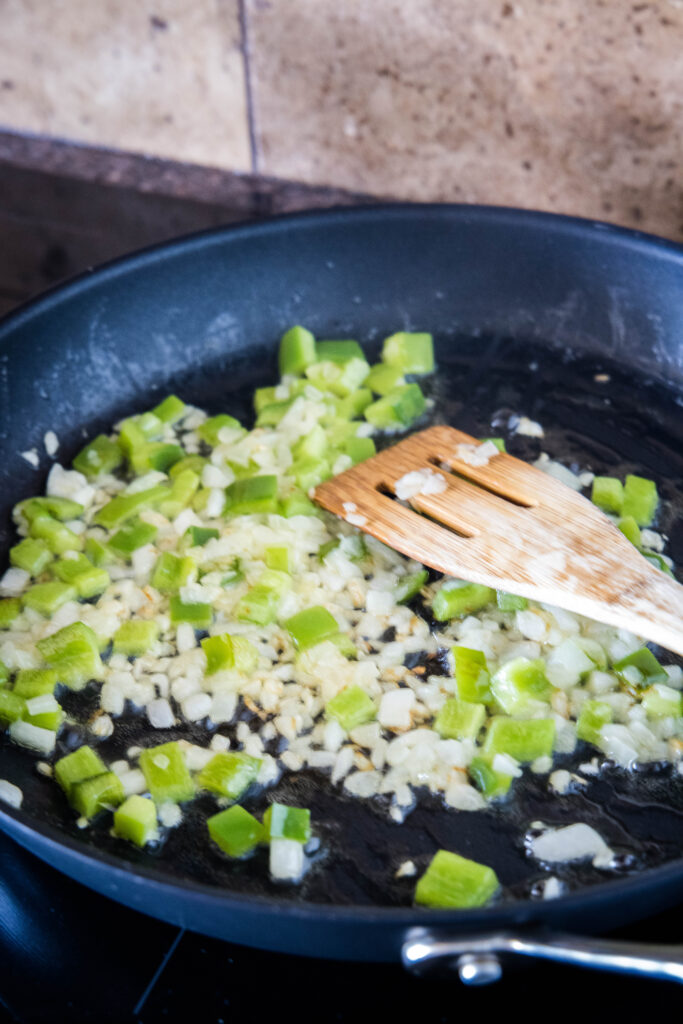 Diced onions and bell pepper sauteing in a large skillet, with a wooden spatula.