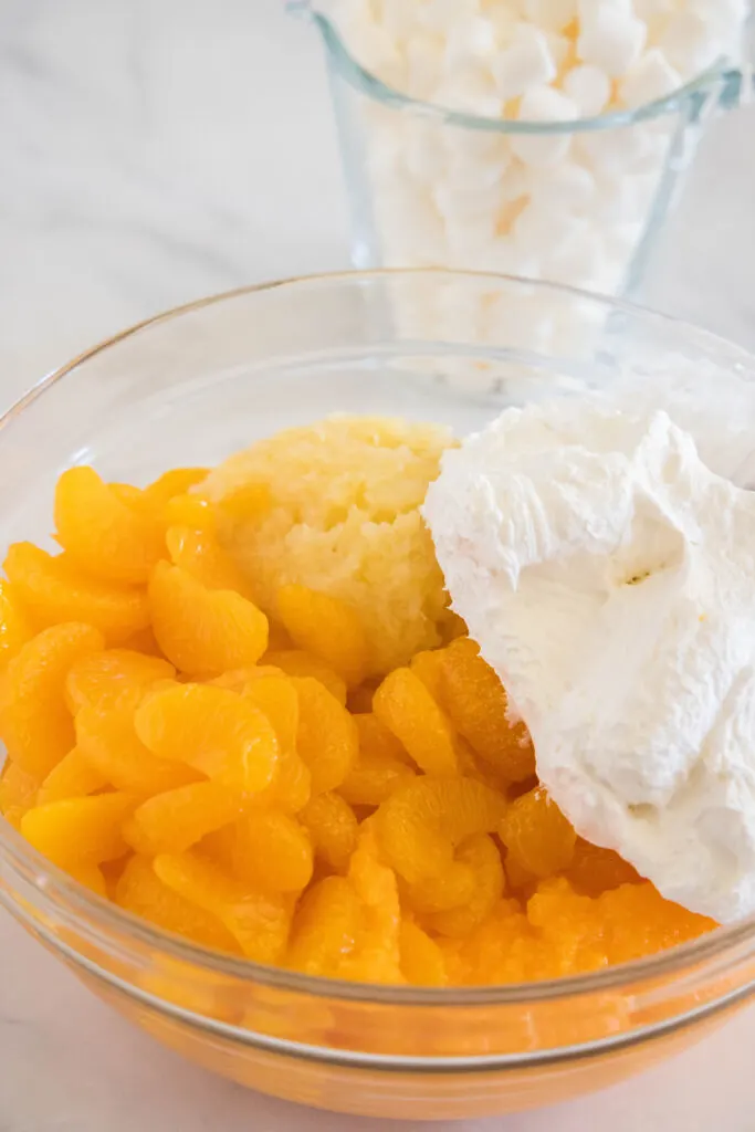 Canned mandarin oranges, crushed pineapple, and Cool Whip added to a glass bowl.