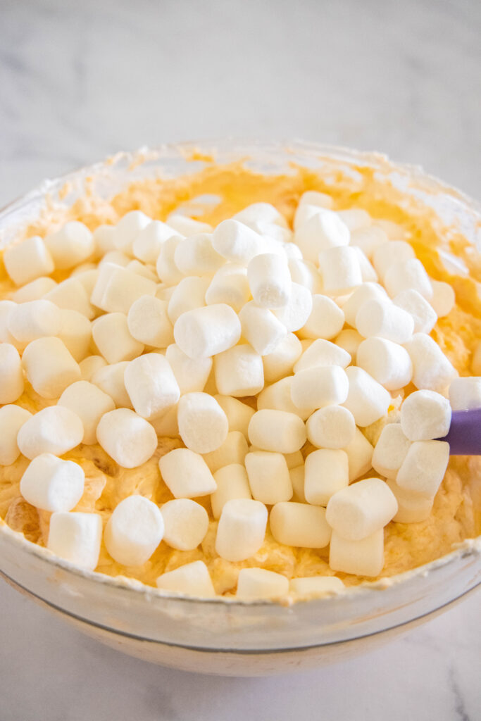 Marshmallows added to orange fluff in a glass bowl.