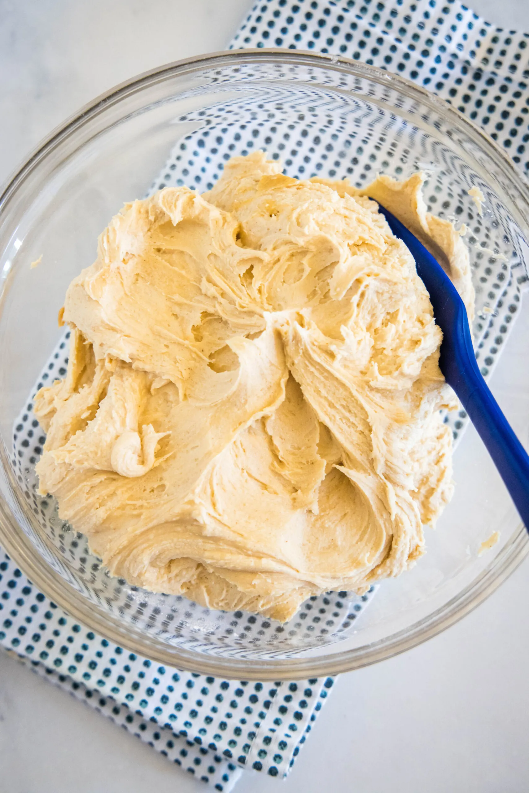 Overhead view of peanut butter frosting in a glass bowl with a blue stirring spoon.