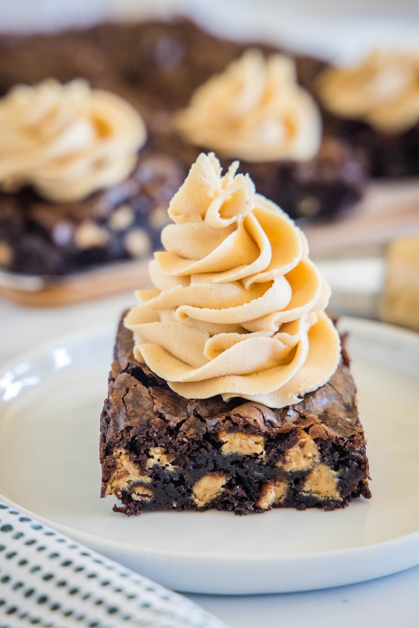 A chocolate brownie topped with a swirl of peanut butter frosting on a plate, with more frosted brownies in the background.