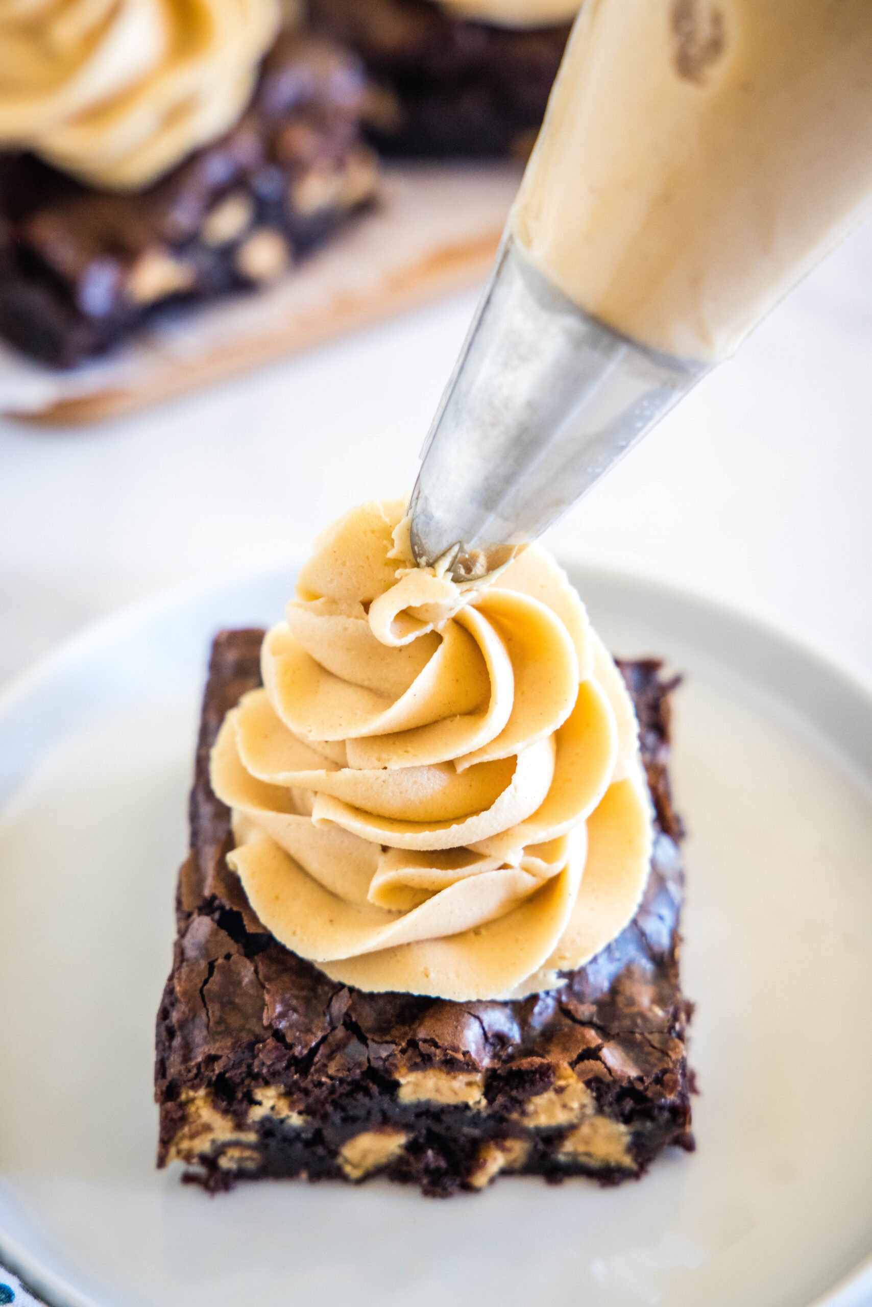 The tip of a piping bag piping a swirl of peanut butter frosting over a chocolate brownie on a white plate.