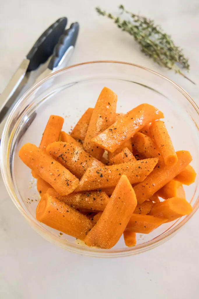 Raw carrots tossed in olive oil and seasoning in a glass bowl.