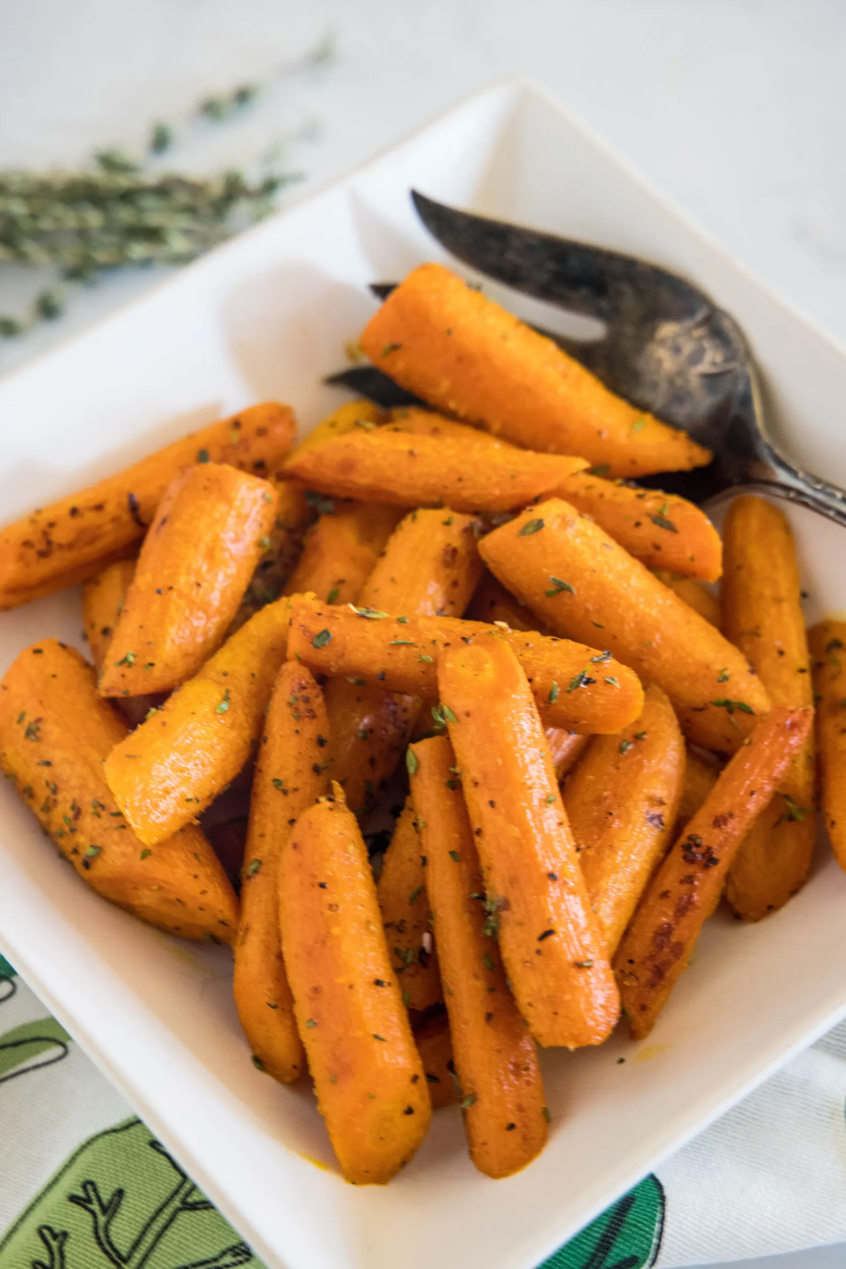 Seasoned roasted carrots in a square white bowl with a serving spoon.