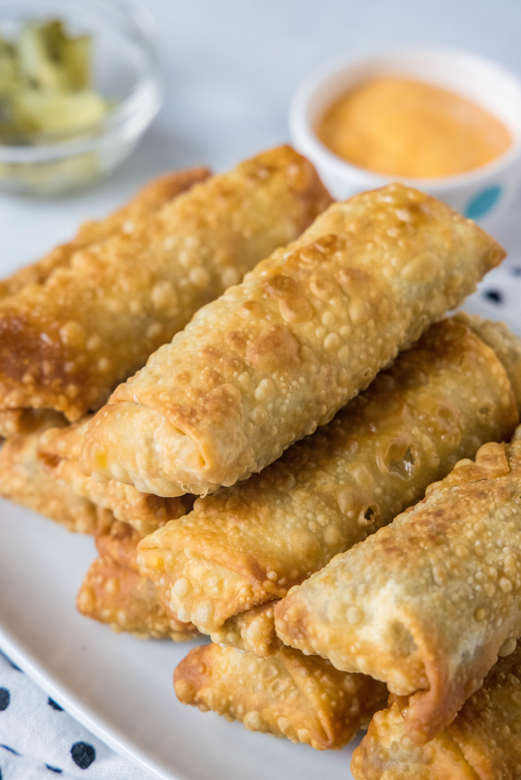 Cheeseburger egg rolls stacked on a platter with a bowl of dipping sauce in the background.