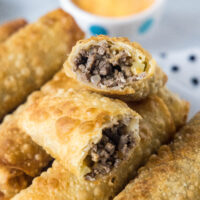 Close up of cheeseburger egg rolls stacked on a platter, with one egg roll broken in two and a bowl of dipping sauce in the background.