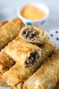 Close up of cheeseburger egg rolls stacked on a platter, with one egg roll broken in two and a bowl of dipping sauce in the background.