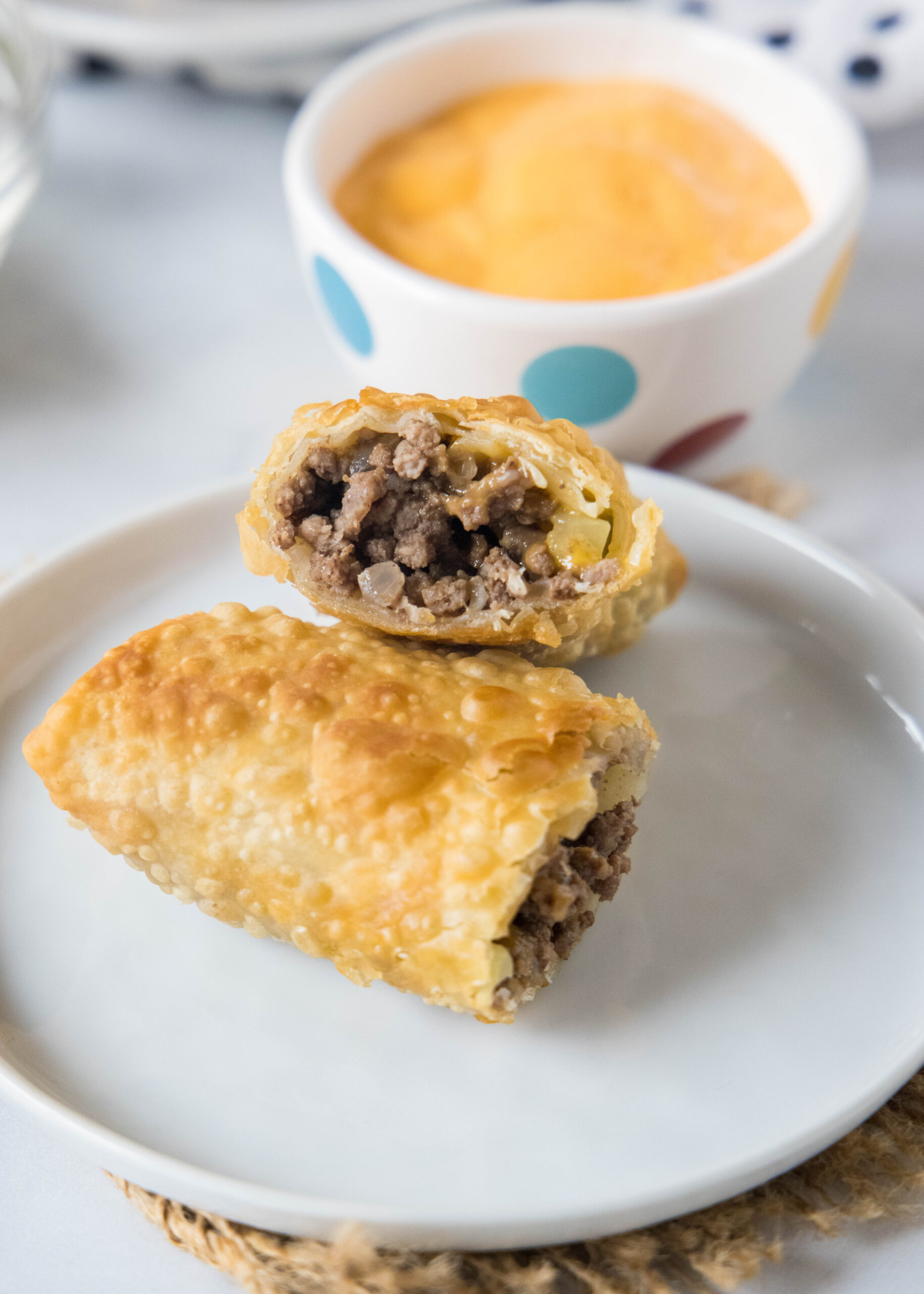 Two halves of a cheeseburger egg roll stacked on a white plate with a bowl of dipping sauce in the background.