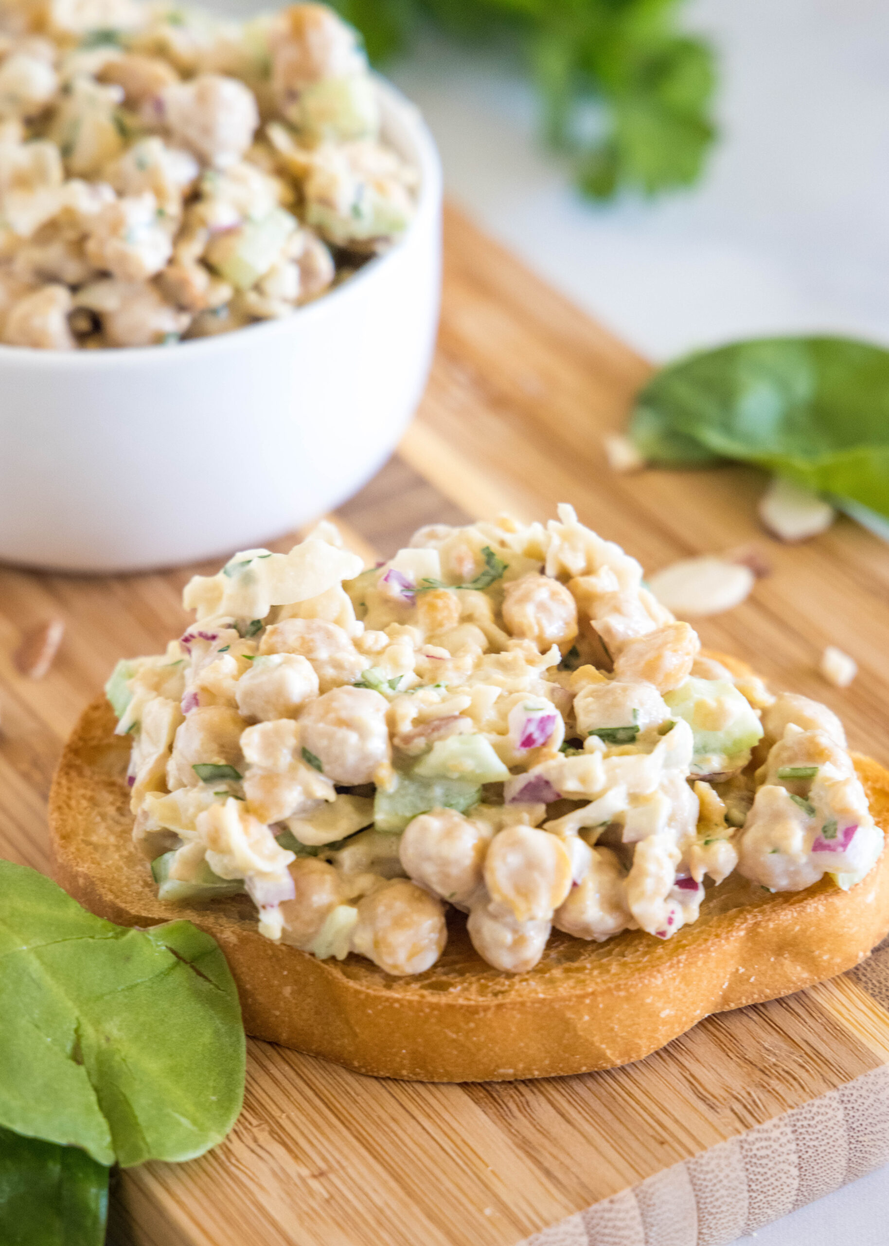 Chickpea chicken salad served on a slice of bread on a wooden cutting board, with a bowl of chickpea salad in the background.