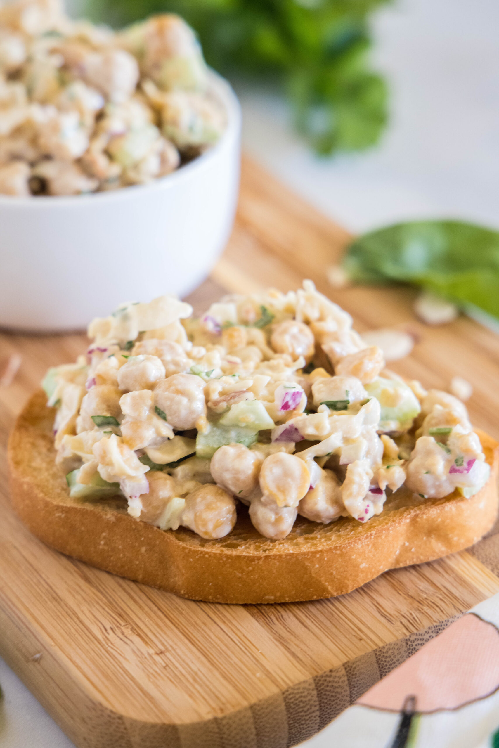 Chickpea chicken salad served on a slice of bread on a wooden cutting board, with a bowl of chickpea salad in the background.