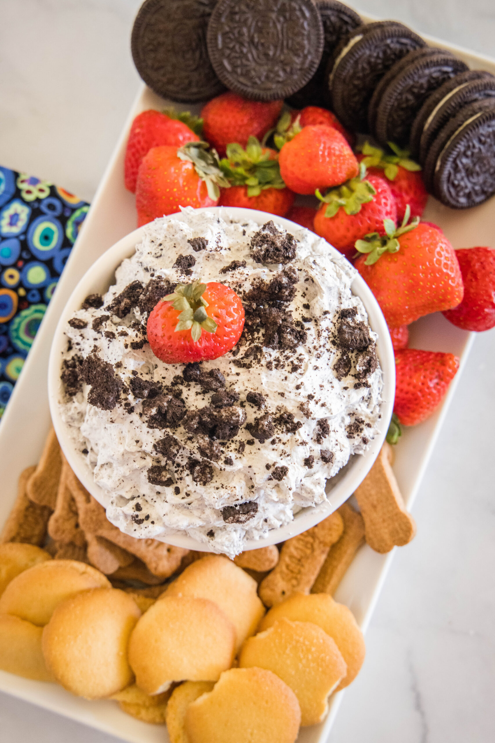 Overhead view of a bowl of Oreo fluff topped with crushed Oreos and a strawberry, surrounded by a platter of dippables.